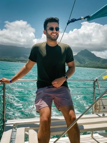 Prateek standing on a sailboat deck with green mountains and blue water behind him.